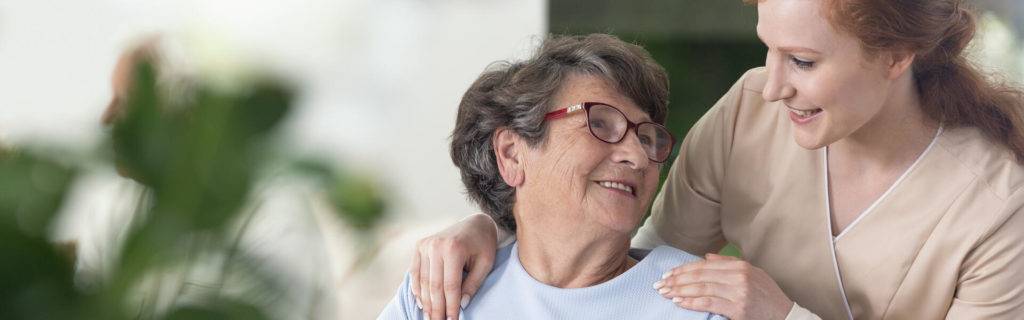 Close-up of a tender caregiver with her hands on the shoulders of a senior woman inside her home. Blurred surrounding. Panorama.